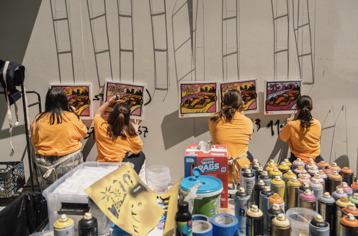 Four participants in yellow shirts creating colorful spray-paint artwork of race cars against a city skyline at an outdoor workshop, with spray cans and stencils on the table.