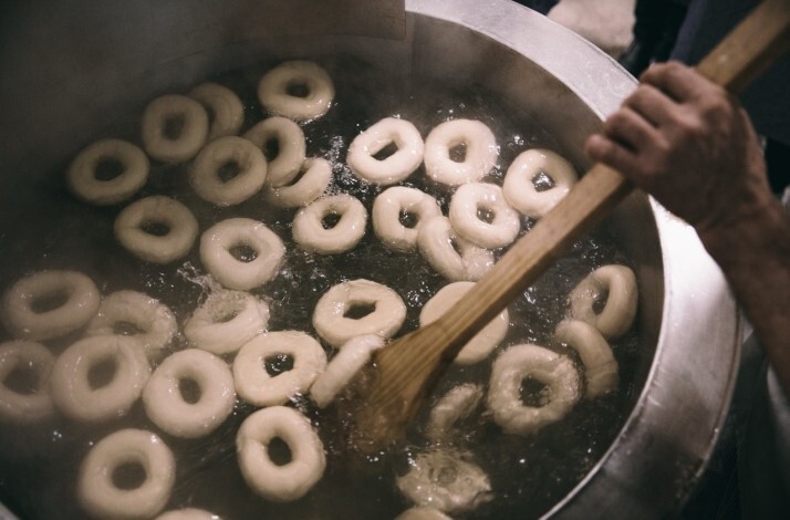 : Dozens of bagels boiling in a large commercial kettle, being stirred with a wooden paddle at Russ & Daughters.