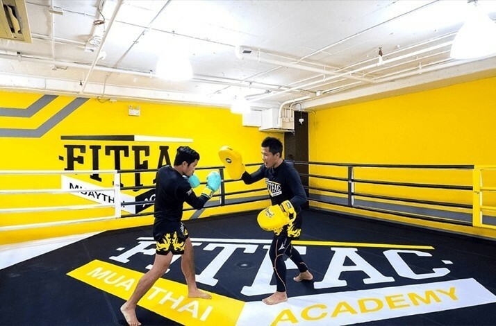 A Muay Thai student practices a kick with pads in a bright training gym.