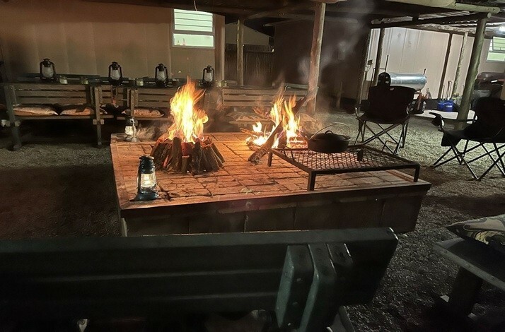 A campfire is burning in a rustic outdoor setting, surrounded by wooden benches, foldable seats and camping equipment at Kruger National Park