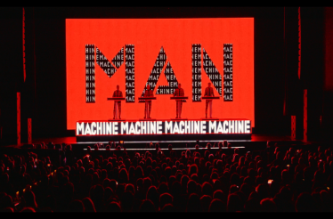 Kraftwerk performing live with red backdrops spelling MAN MACHINE in front of a seated audience.