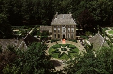An aerial view of Chateauhotel de Havixhorst, featuring a circular flowerbed design garden situated in front of the hotel's entrance. The entrance is surrounded by various large estate with other gardens, and an orange carpet leads to the entrance of the hotel.