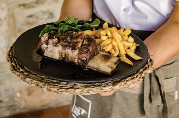 Server holding a black plate with grilled ribs, chips, and greens at Habitat Cigueña Negra restaurant.