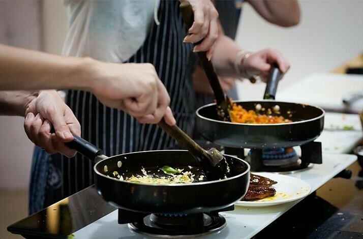 Two people are cooking in a kitchen of Studio Cooking Class, stirring food in frying pans on a stove.