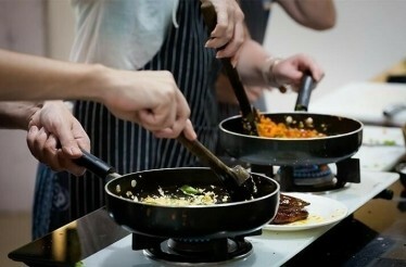 Two people are cooking in a kitchen of Studio Cooking Class, stirring food in frying pans on a stove.