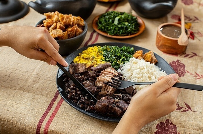 A person is seen cutting into a plate of Feijoada, a traditional Brazilian dish with black beans, rice, and pork.