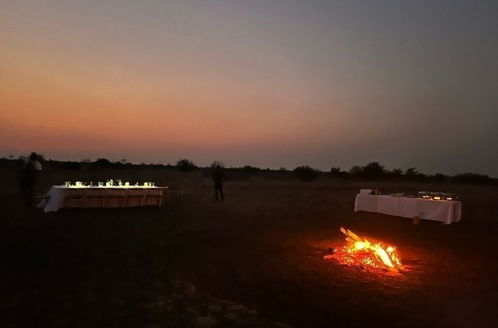 A campfire glows at sunset near two tables set for dinner in the open wilderness