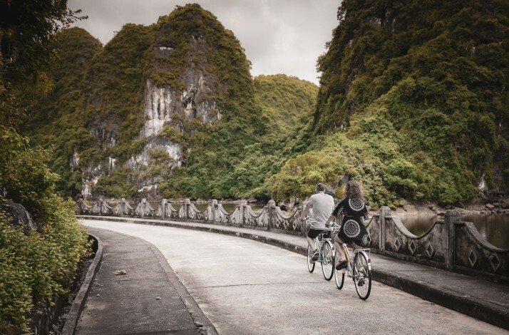 A couple is seen riding tandem bicycles on a road in Hạ Long Bay's Heritage Line surrounded by lush greenery and rocky hills.