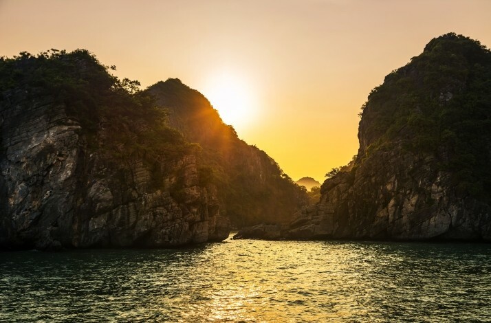 A serene seascape at sunset, showcasing a tranquil bay between towering limestone cliffs at Hạ Long Bay's Heritage Line.