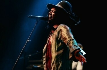 Fantastic Negrito in vibrant attire and a wide-brimmed hat passionately sings on stage under dramatic lighting