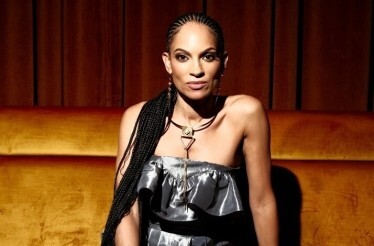 Promotional banner for Goapele in a silver ruffled top and gold necklace sits on a velvet couch.
