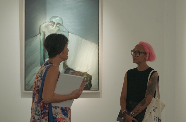 Angeline Gong and an art enthusiast discuss paintings with dark, emotive imagery displayed on minimalist white gallery walls