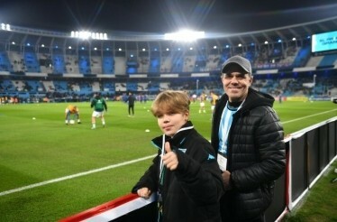 A child showing thumbs up to the camera with football prod warming up in the background