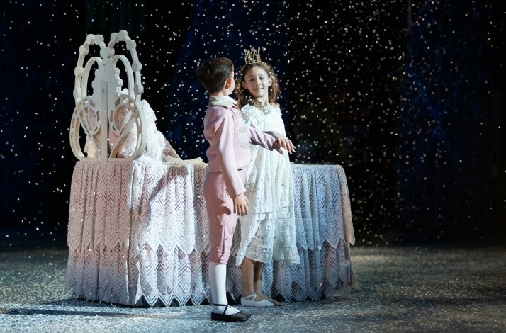 Young Clara and the Prince on stage in a snowy scene with a decorative bed.