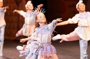A smiling young Clara in pastel costume dances joyfully in a group performance.