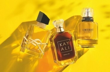 A close-up displays three perfume bottles of varying colors and designs, positioned on a bright yellow background. 