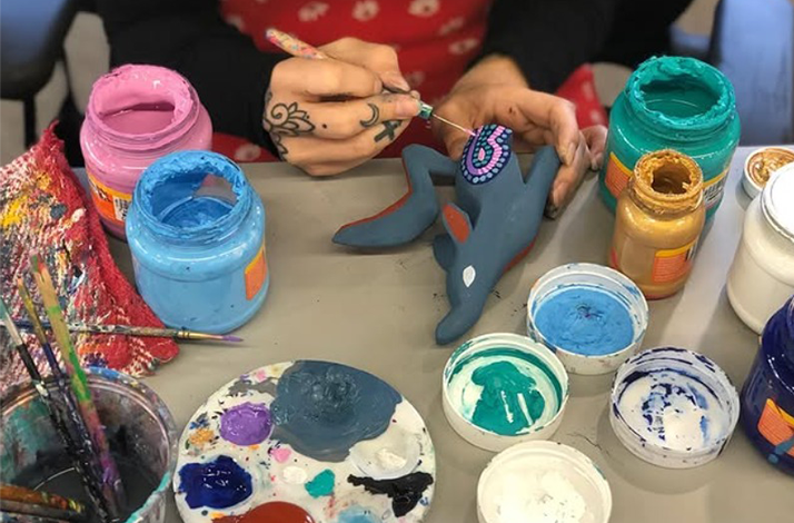 The artist is painting a small gray figurine with blue and pink dots.