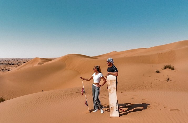Two people holding sandboards, standing on sand dunes under a clear blue sky.