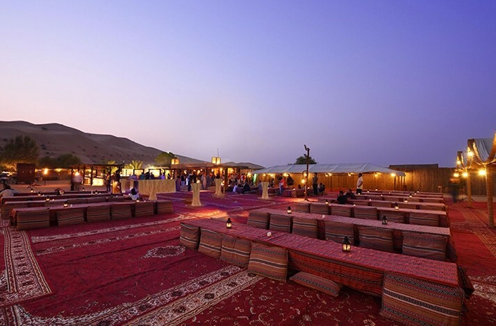 Desert camp setup with traditional seating, rugs, and ambient lighting at sunset.
