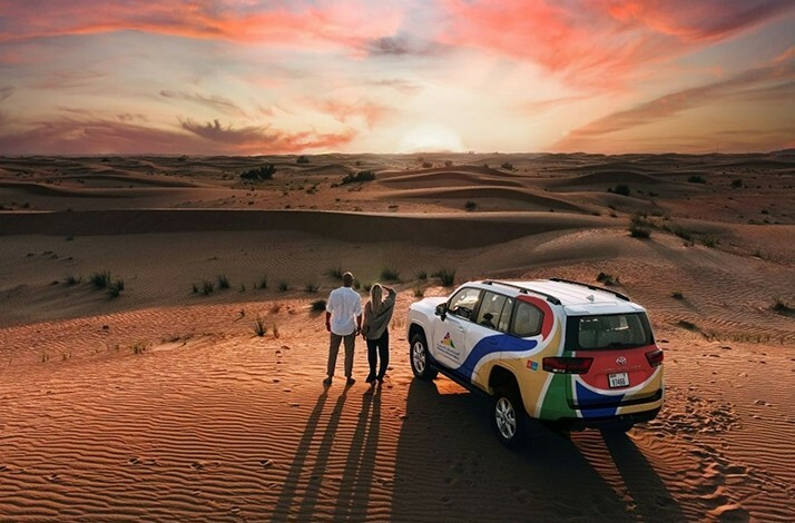 An SUV parked in the Dubai Desert Conservation Reserve as people admire the sunset over sand dunes.