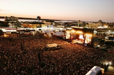 Large crowd gathers at an outdoor music festival by the waterfront, with a brightly lit stage at sunset.
