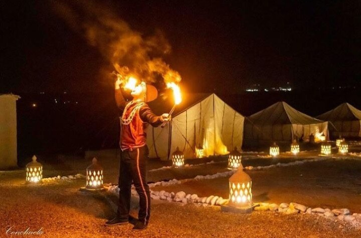 A nighttime scene at Agafay Desert captures a performer breathing fire amidst illuminated lanterns and tents.