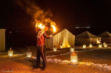 A nighttime scene at Agafay Desert captures a performer breathing fire amidst illuminated lanterns and tents.