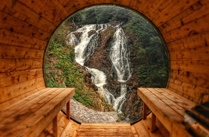 A view of a cascading waterfall framed by a wooden sauna's circular window, at Calumet Falls