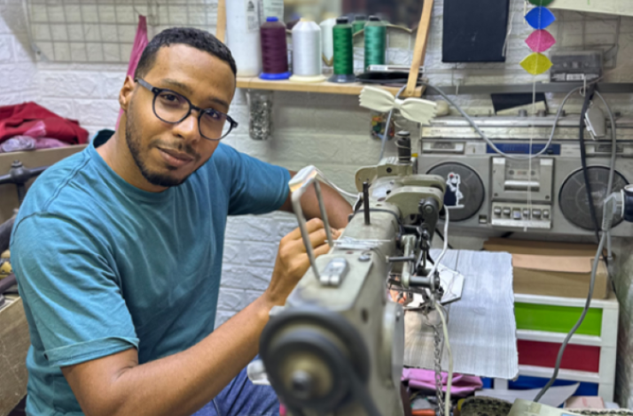 A man in a blue shirt operates a sewing machine, focused on his task.