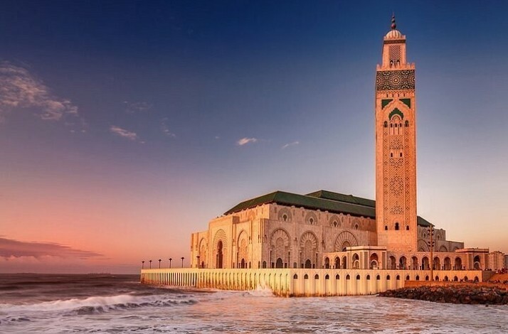 Hassan II Mosque stands majestically by the ocean, bathed in warm sunset light and waves below.