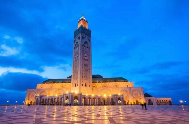 Outside view of Hassan II Mosque illuminated at dusk, with its grand minaret and patterned courtyard under a blue sky.