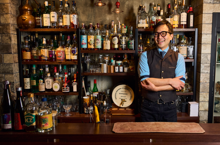 Smiling bartender in a vest and glasses standing with arms crossed in front of shelves stocked with an extensive whiskey and spirits collection.