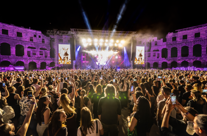 A vibrant night concert scene inside the Pula Arena in Pula, Croatia, showing a dense crowd of people with their phones raised.
