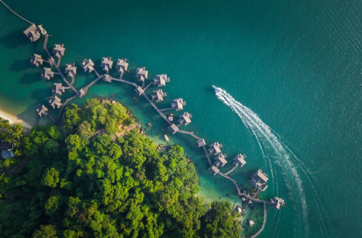 Aerial view of overwater villas, green island, and a boat on turquoise waters at Pangkor Laut Resort.