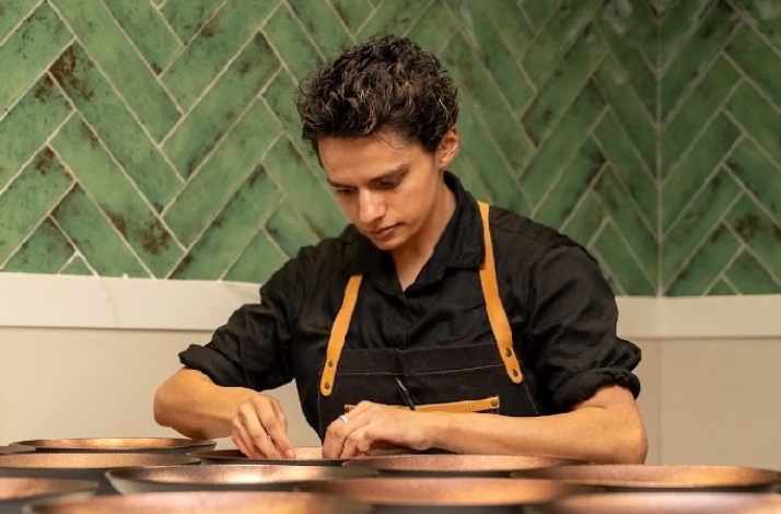 A chef in a black shirt and apron carefully plating dishes at a counter against a green herringbone-tiled wall.
