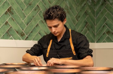 A chef in a black shirt and apron carefully plating dishes at a counter against a green herringbone-tiled wall.