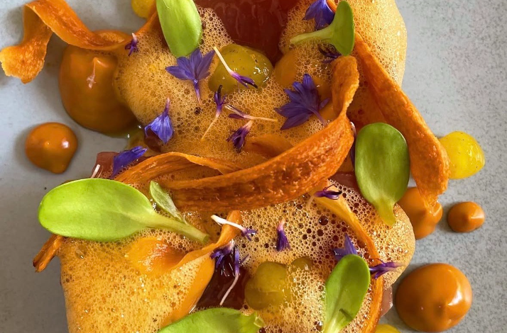 A close-up of an elegantly plated dish garnished with edible purple flowers, microgreens, and orange foam.