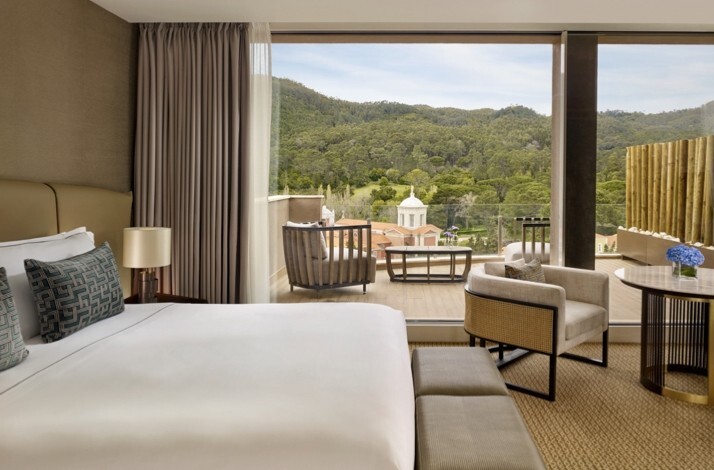 Elegant hotel room at Penha Longa Resort, containing a large bed and seating area, opening onto a balcony with scenic views of forested hills and a historic chapel.