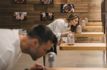 Two visitors leaning over tiered wooden shelves to smell glass-domed aroma samples in a sensory exhibition room at The Wine Experience - Wine Museum.