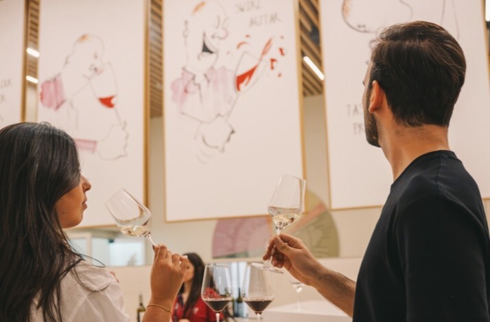 Two visitors smelling white wine during a guided tasting session at The Wine Experience - Wine Museum, with illustrated artworks on the gallery walls.