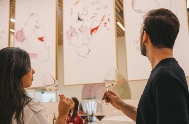 Two visitors smelling white wine during a guided tasting session at The Wine Experience - Wine Museum, with illustrated artworks on the gallery walls.