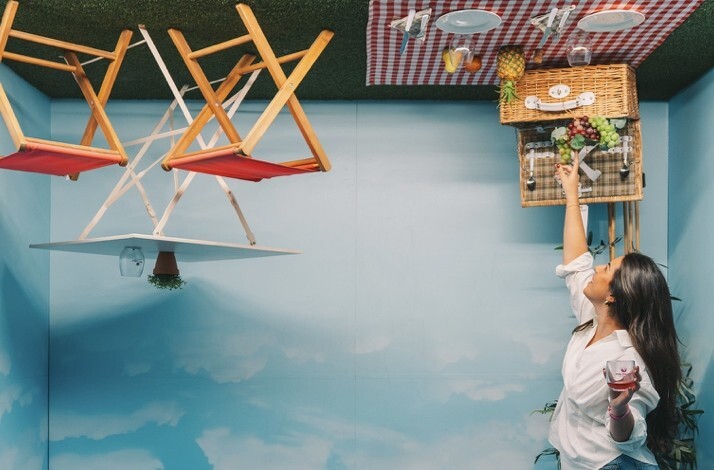 Visitor reaching toward an inverted picnic installation inside a The Wine Experience - Wine Museum gallery, with suspended basket and chairs overhead.