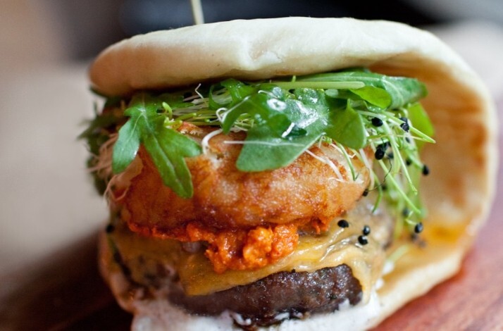 Bäco sandwich from le dräQ, layered with beef patty, red harissa, melted cheese, crispy onion, microgreens, and pea shoots.