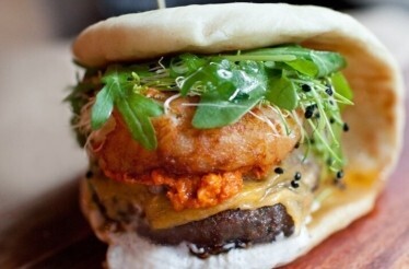 Bäco sandwich from le dräQ, layered with beef patty, red harissa, melted cheese, crispy onion, microgreens, and pea shoots.