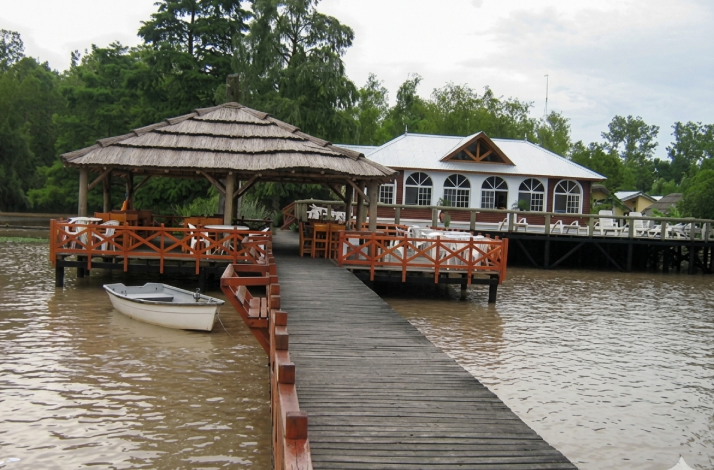 Wooden dock leading to a thatched-roof gazebo and a riverside lodge, with a small rowboat moored at the edge.