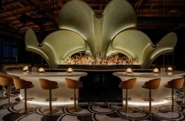 Grand curved bar at Lucia LA topped by an illuminated petal-shaped installation above a full spirits display.