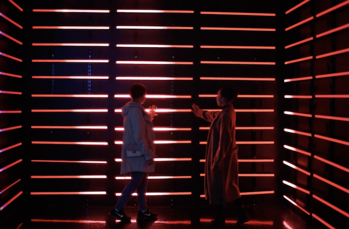 Two people facing each other with hands raised beside walls of red neon strip lights in a darkened room.