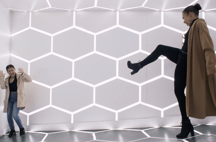Two visitors laughing and posing beside a floor-to-ceiling illuminated hexagonal light wall.