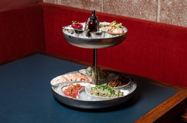 Two-tiered seafood platter on ice, featuring shrimp, oysters, cev iche, and garnishes, set on a dark blue table with a red booth, served at Loreto.