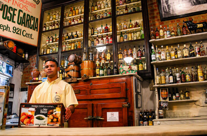 A bartender standing behind a counter in a rustic bar with shelves filled with liquor bottles in Rio.